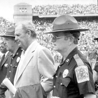 Alabama coach Bear Bryant is escorted by Alabama State Troopers off the field at Memorial Stadium on Sept. 17, 1977, after his fourth-ranked Crimson Tide lost to Nebraska 31-24.
