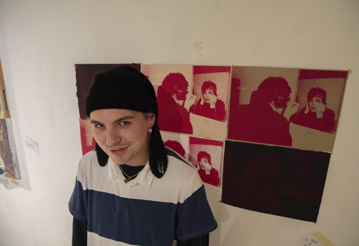 Junior studio art major Ethan Justice poses next to his print “Guyliner” made to represent a different view of masculinity and what it means to “be a man” in the Fine Arts Center's Cube Gallery on Oct. 15, 2024.